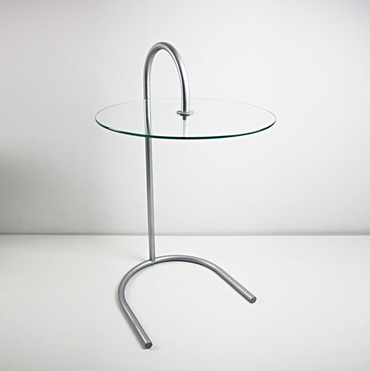 A modern side table with a clear glass circular tabletop and a curved silver metal base, photographed against a plain light gray studio background. RY table by IKEA designed by Tord Björklund