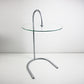 A modern side table with a clear glass circular tabletop and a curved silver metal base, photographed against a plain light gray studio background. RY table by IKEA designed by Tord Björklund