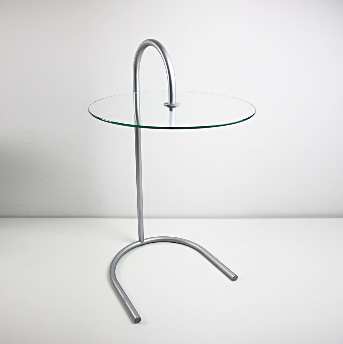 A modern side table with a clear glass circular tabletop and a curved silver metal base, photographed against a plain light gray studio background. RY table by IKEA designed by Tord Björklund
