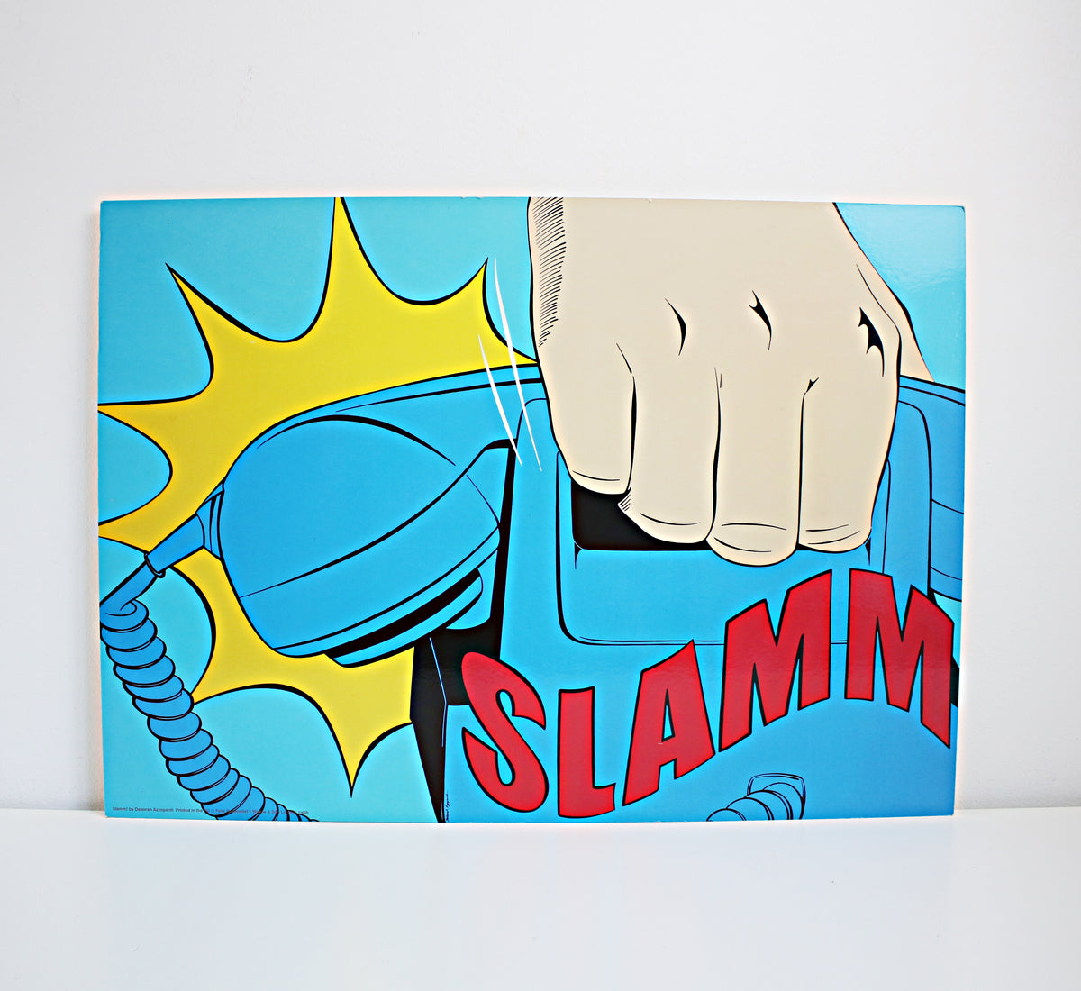 A square pop-art style print or poster showing a hand slamming a blue telephone receiver, with a yellow comic burst and the word "SLAMM" in red, photographed against a light gray wall and white surface. IKEA 1990s art. Vintage